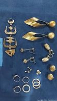 Overview photo of all jewelry items laid out on a surface showing fish pendant, leaf-shaped gold plated earrings, gold plated heart studs, silver pairs of earrings and rings.