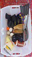 Top view of all sporting equipment in a clear plastic bin including badminton racquets, diving booties, baseball gloves and balls, football, and protective gear.