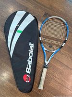 Tennis racket with cover on a wooden floor showing full racket and black cover with Babolat logo.