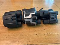 Three compact binoculars displayed side by side on wooden surface, showing size and design variation.