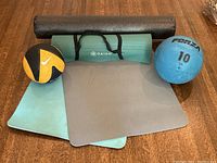 Photo showing two yoga mats, one green Gaiam mat with carry strap and one grey mat, two medicine balls (blue Forza 10 lb and black/yellow Nike 6 lb), and a black foam roller lengthwise behind the mats and balls.