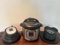 Front view of Instant Pot electric pressure cooker with air fryer lid and basket on sides shown on wooden surface against white wall.