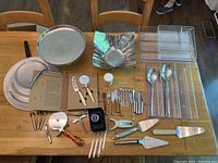 Full view of all kitchen utensils arranged on a table including pizza pans, pizza cutter set in box, pie servers, corkscrew, forks, knives, and utensil organizer tray