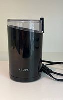 Front and side view of the black Krups coffee grinder with clear lid on top and power cord wrapped beside it.
