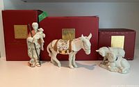 Three Lenox porcelain figurines displayed side by side with their original red and gold Lennox boxes behind them. Figures include shepherd boy with lamb, standing donkey, and pair of sheep.