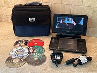 Panasonic portable DVD player opened with screen displaying a movie scene, showing included DVDs, Roots branded carrying case, and power adapters arranged on a tile floor.
