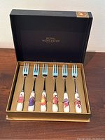 Full view of set in black Royal Worcester box with gold interior showing six porcelain handle pastry forks with fruit images and metal tines