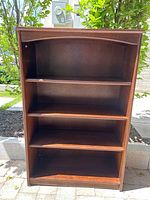 Front view of the solid wood bookshelf showing three adjustable shelves and the bottom shelf, placed outdoors with greenery in the background.