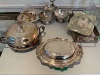Full view of vintage silver plated serving dish lot including various covered dishes, bowls, and trays arranged on table.
