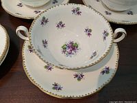 Close-up of a two-handled soup bowl resting on a matching saucer showing purple violet floral decoration and gold trim detailing.