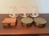 Three hand blown whiskey glasses with etched numbers 5, 1, and 6, two stacks of brass coasters, and a wooden box labeled iiiMY whiskey stones.