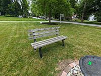 Photo of one weathered wooden outdoor bench on grass with black metal legs and three wooden slats on the back.