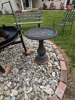 Cast aluminum bird bath on rocky ground near lawn and patio furniture, showing weathered texture and condition