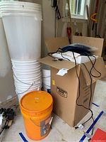 Photo of stacked white 5-gallon buckets, one orange bucket, and a box with electrical cords resting on it in a garage.