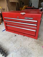 Red Craftsman metal tool chest with four drawers, placed on a garage floor with various items around. Shows overall condition and placement.