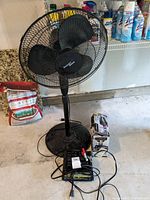 Front view of the Nantucket Breeze standing fan with charger and cables on floor near it. Fan is black with round base and metal grill.