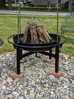 Fire pit shown outdoors on rock base with stacked logs inside, featuring rotisserie skewers and crank handle mounted on tripod legs.