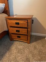 Solid wood night stand positioned next to bed on carpeted floor. Three drawers with metal handles, top drawer slightly ajar revealing drawer rail.