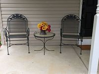 Front view of two black metal floral motif patio chairs with a round glass top metal table between them, placed on a porch.