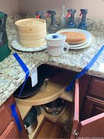 Photo shows countertop with bamboo steamers stacked on a white egg plate, several spray bottles arranged behind, a stack of platters and tortilla warmer. Corner cabinet doors open showing pots and pans inside on two rotating shelves.