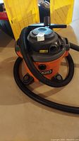 Full view of the orange and black Rigid shop vacuum cleaner with hose attached and surrounding items.