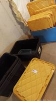 Four black plastic storage boxes stacked with yellow lids next to a blue plastic tote with yellow lids in a basement corner on a concrete floor