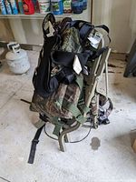 Folded tree stand featuring a camouflage padded seat and backrest with multiple straps and harness attached, resting on a concrete floor in a garage.