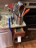 Two stainless steel utensil holders filled with a variety of kitchen utensils on a kitchen counter, visible drawers beneath containing more utensils, a black handheld sharpener on the counter.