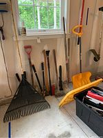 Wide view of the yard tools including rakes, shovels, and clippers leaning against a wall in a garage near a window.