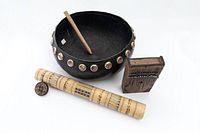 Three tribal items arranged: black rattle amplifier with bottle caps, kalimba thumb piano, and decorated bamboo container with lid.