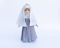 Full view of the nun doll dressed in a gray habit with a white head cover, white shoes, and socks, standing upright on a stand.