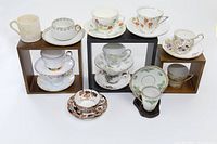 Set of 10 fine china cups and saucers with floral and decorative patterns displayed in wooden cube stands, plus two small mugs, one white with brown-gold pattern and the other one partly visible.