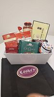 Cooke's branded white basket filled with various gourmet food items such as pasta sauce, caramel bites, tea, jams and nuts placed on a surface alongside a Cooke's black reusable shopping bag.