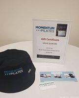 Gift certificate, black bucket hat with Momentum Pilates logo, and business cards displayed on white table.