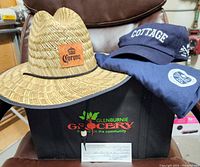 Photo showing the Glenburnie Grocery insulated grocery tote bag filled with a blue Cottage Springs T-shirt, matching ball cap, and a woven Corona sun hat on top, plus visible $50 gift certificate in front