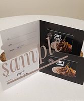 Photo showing two folded black holders each containing a One Eyed Jack Pub and Grill gift card with images of grilled meat and vegetables. The holders have handwritten amounts of $75 and $100 respectively.