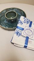 Photo showing a glazed blue-green pottery serving dish with a matching small bowl inside, alongside a set of three neatly folded white cotton tea towels with blue floral Buckingham Palace designs, tied with blue ribbon and tag.
