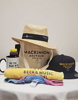 Photo of MacKinnon Brothers Brewing Co. items including straw sun hat, black ballcap, tie-dyed t-shirt, canvas tote, Erin's Hot Honey, Lodge Coffee House mug.
