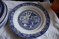 Close-up photo of one Blue Willow style plate showing blue pastoral pattern with boats, trees, pagoda, bridge, and floral border.