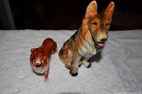Both porcelain dog figurines placed on white fabric; one sitting German Shepherd and one standing Collie.