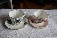 Full view of two teacups and saucers on a white fabric surface, one with blue floral pattern and one with pink floral design.
