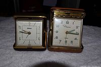 Two vintage travel alarm clocks with gold-tone and leather casing, placed side by side on a white towel.