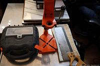 Overhead photo showing black mouse sander with carrying case, bright orange drill press stand, and metal miter saw guide with wooden handle on a table.