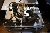 Seven antique mechanical tools on metal surface, including two apple peelers, cherry pitter, meat grinder, oil can, and two wood planers.