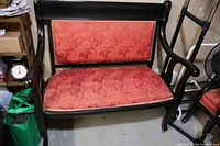 Front view of the black painted wooden Edwardian settee with faded red floral upholstery and curved armrests, situated in a storage-like setting.