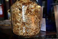Close-up photo of a glass jar filled with crinkled gold metallic leaf flakes, showing texture and shine of the flakes inside.