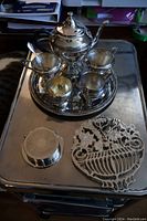 Photo showing all silver plate items together including tea pot, cream and sugar sets, tray, spoon, trivet, and coasters on a surface.