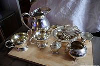Silver-plated assorted tableware including milk and sugar bowl set, water jug, small creamer, covered container, and fish forks displayed on and around a wooden table surface.