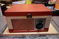 Closed view of the red and cream textured portable record player with metal latch on top, resting on a wooden board. Shows front grille and control area.