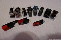 Top view of 11 vintage turntable cartridges arranged in a row, showing different shapes, colors, and styles including black, red, and silver plastic housings with metal connectors and some needles visible.